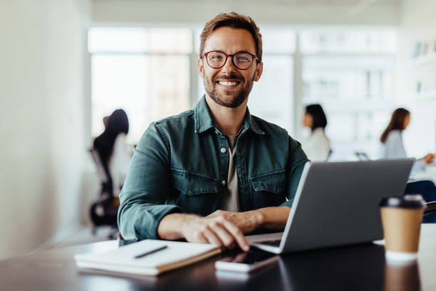 Portrait of a business man sitting in an office with his colleagues in the background. Happy business man working in a co-working office.